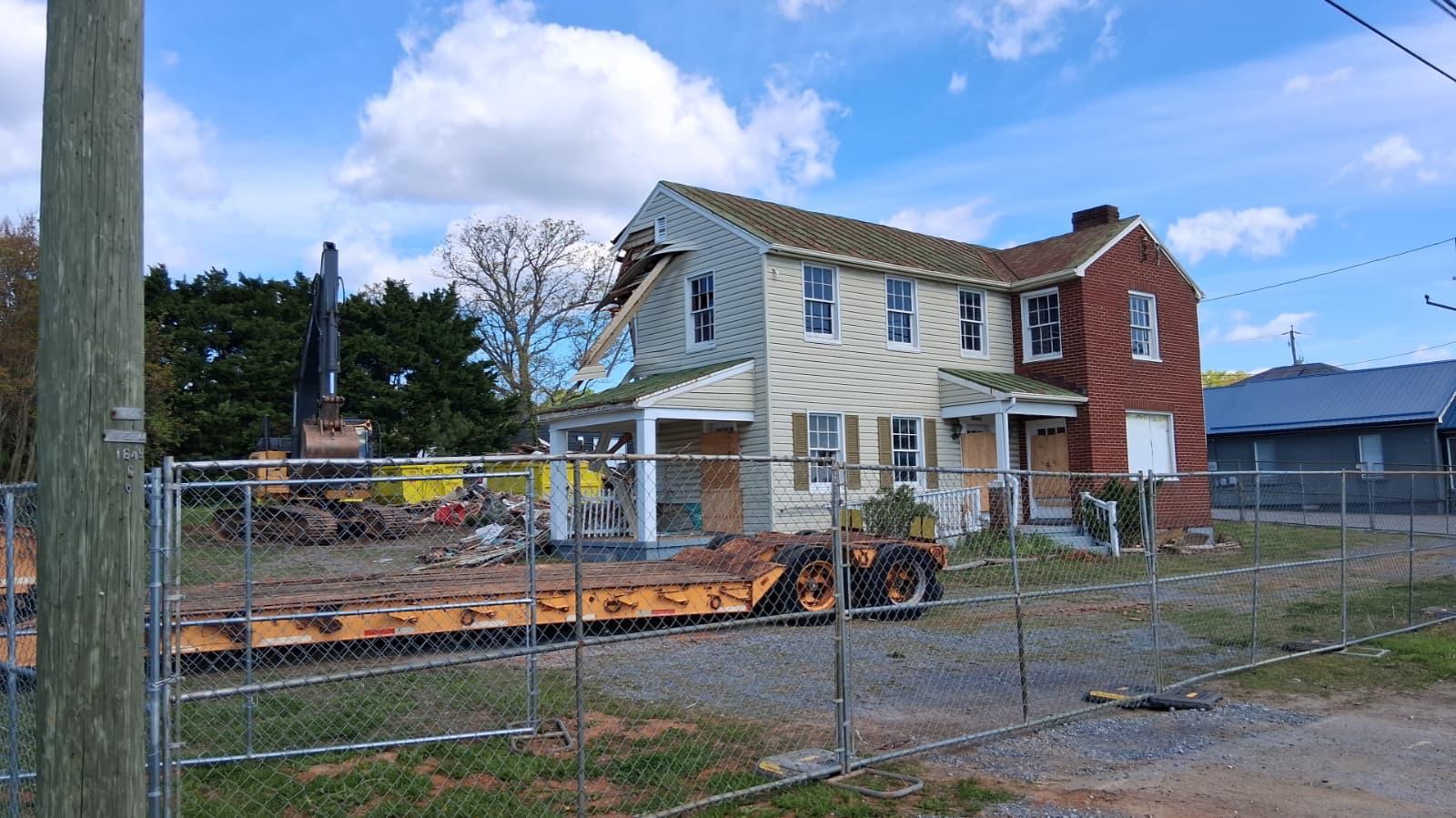 Excavator tearing off the roof and gable of a house during a structure demolition, dumpster staged in the yard