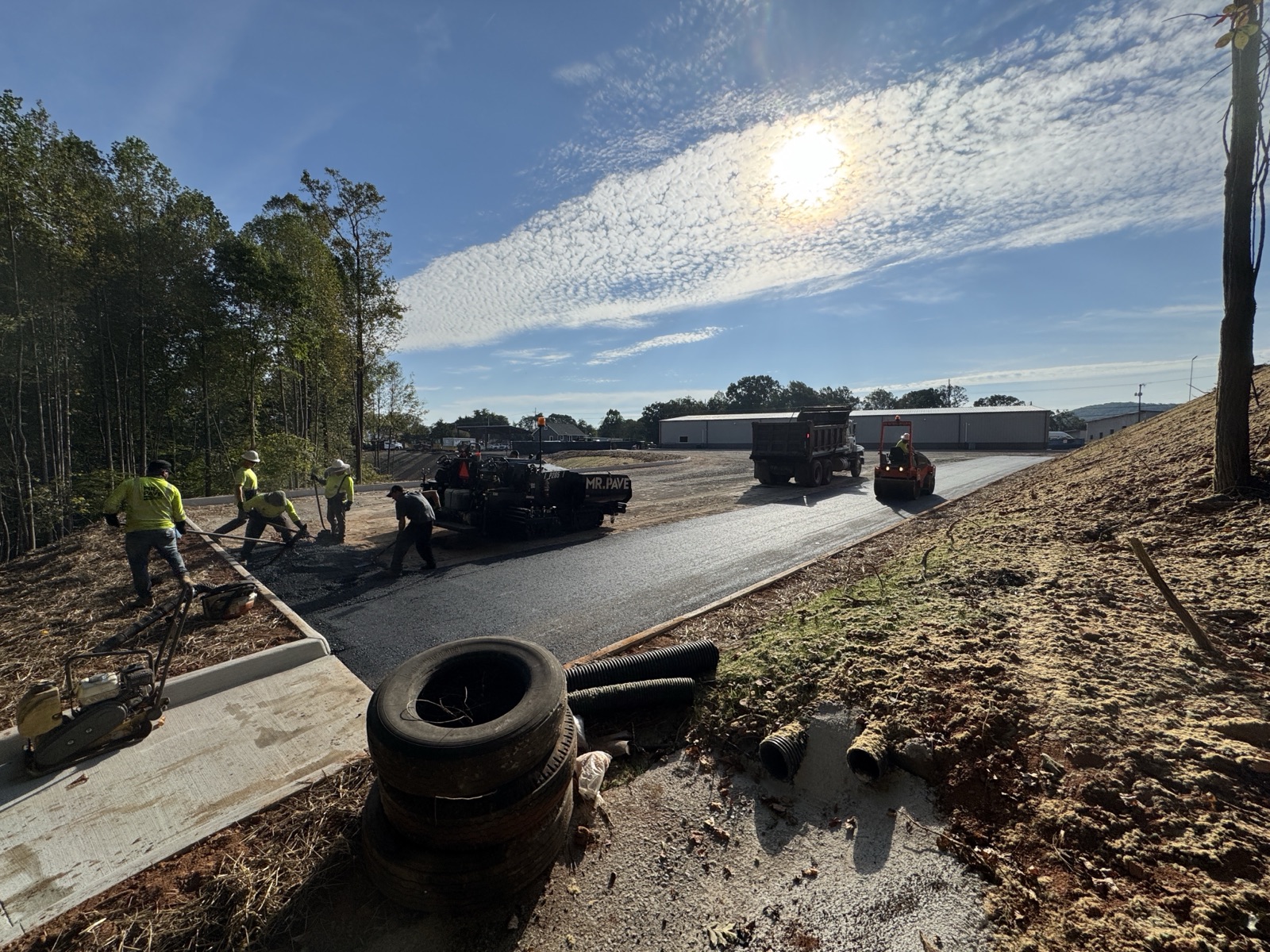Mr. Pave crew paving a road with a paver, dump truck, and roller staged on site